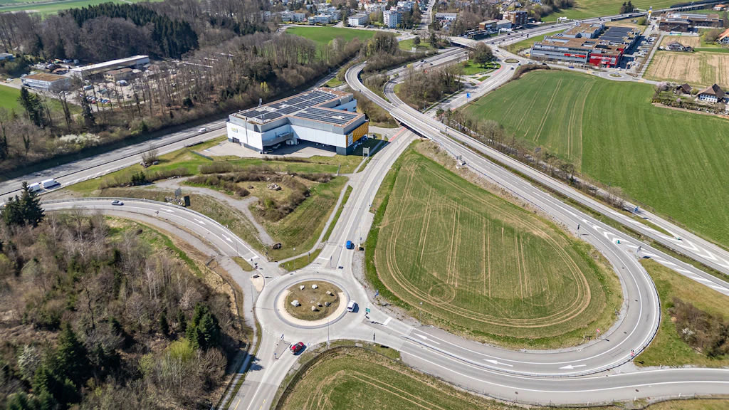Luftaufnahme eines Kreisverkehrs mit angrenzenden Strassen, Feldern und Gebäuden in einer ländlichen Umgebung
