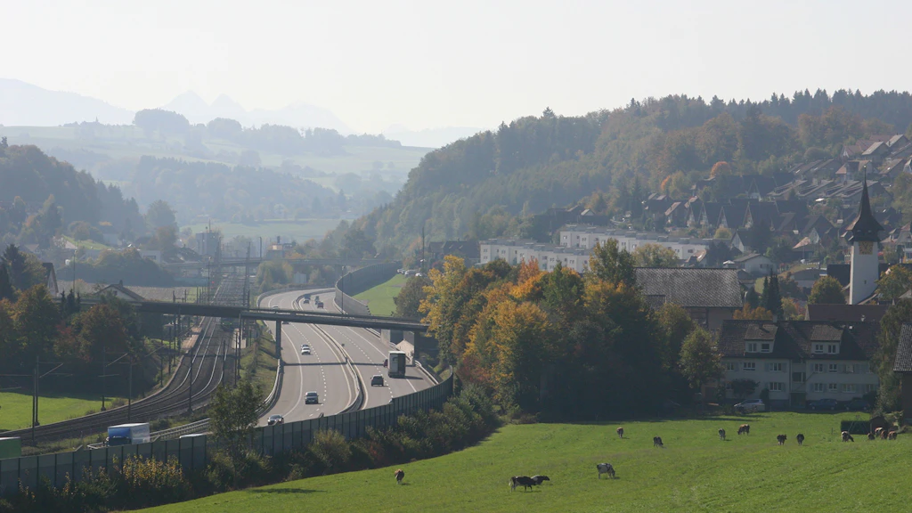 Blick auf eine Autobahn mit vier Fahrspuren und mehreren Fahrzeugen, daneben eine Bahnstrecke und eine Wiese mit grasenden Kühen, im Hintergrund bewaldete Hügel und ein Dorf mit Kirche