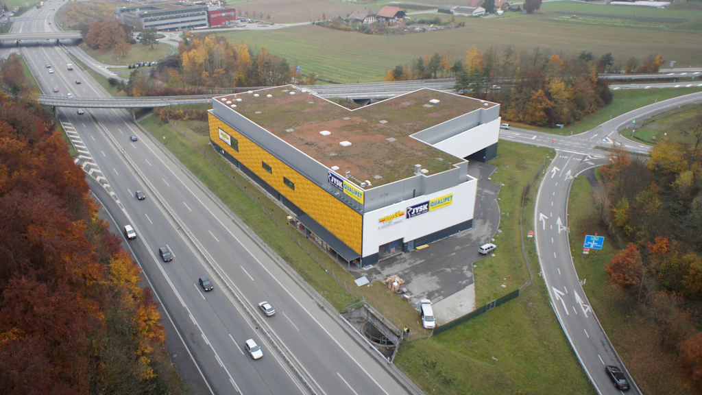Luftaufnahme eines gelb-weisen Gebäudes mit begrüntem Dach neben einer Autobahn und einer angrenzenden Straße mit Kreisverkehr in herbstlicher Landschaft