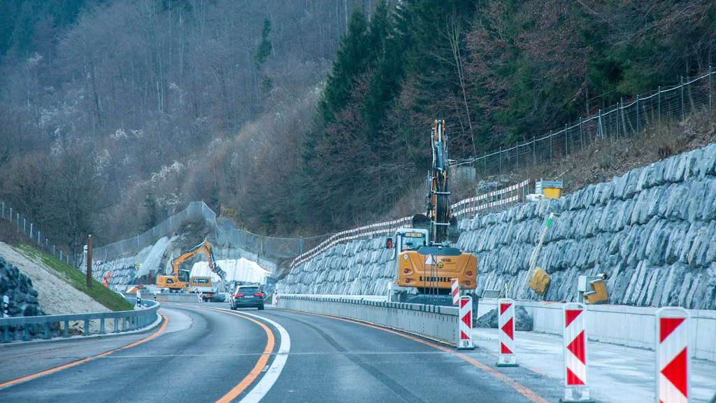 Rue avec un chantier et un mur de soutènement en grosses pierres sur un versant boisé