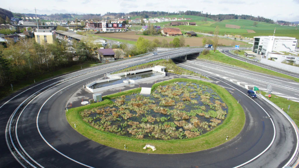 Luftaufnahme einer Kurve einer Autobahnausfahrt mit einem runden Wasserbecken, das mit Schilf bewachsen ist.