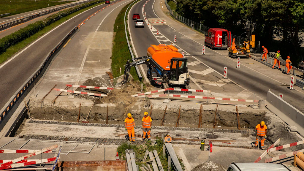Chantier routier sur une autoroute avec plusieurs ouvriers en tenue orange et une machine orange creusant une tranchée encadrée de barrières rouges et blanches