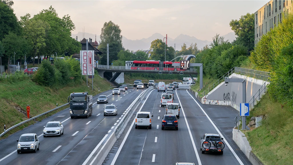Autobahn zwischen Wankdorf und Ostring mit mehreren Fahrzeugen in beide Fahrtrichtungen mit Fahrzeugen auf dem Pannenstreifen bei eingeschalteter Pannenstreifen-Umnutzung. Ein roter Bus fährt über eine Brücke über der Autobahn. Die Autobahn ist von Bäumen und begrünten Böschungen gesäumt, im Hintergrund sind Bäume zu erkennen.