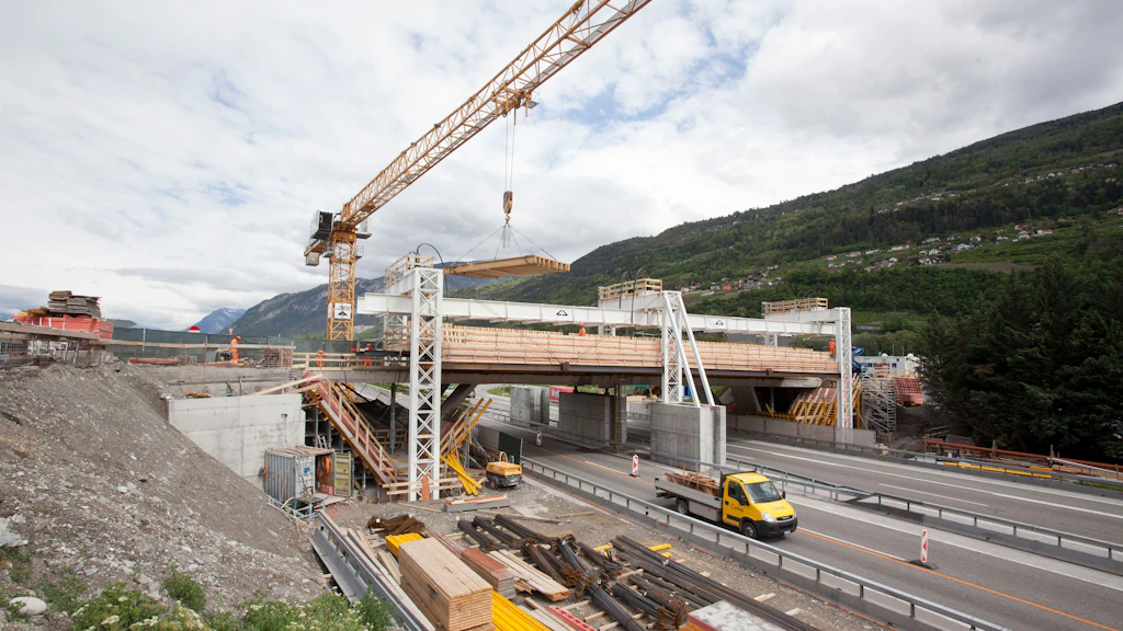 Pont en construction au-dessus d'une autoroute, avec une grue de chantier, des matériaux de construction et une camionnette jaune, entouré de montagnes