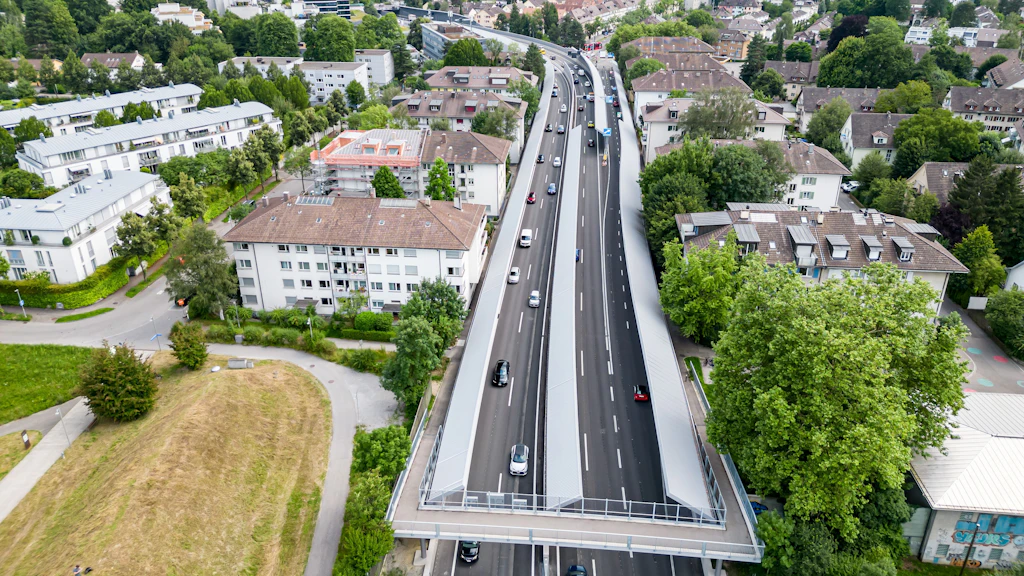 Luftaufnahme der mehrspurigen Autobahn A6 beim Anschluss Ostring. Links und rechts direkt an der Autobahn sind mehrstöckige Wohnblöcke. Zwischen der Autobahn und den Wohnblöcken sind hohe Lärmschutzwände angebracht. Im Vordergrund überquert eine Fussgängerbrücke die Autobahn. Im Hintergrund sind weitere Gebäude und Bäume zu sehen.