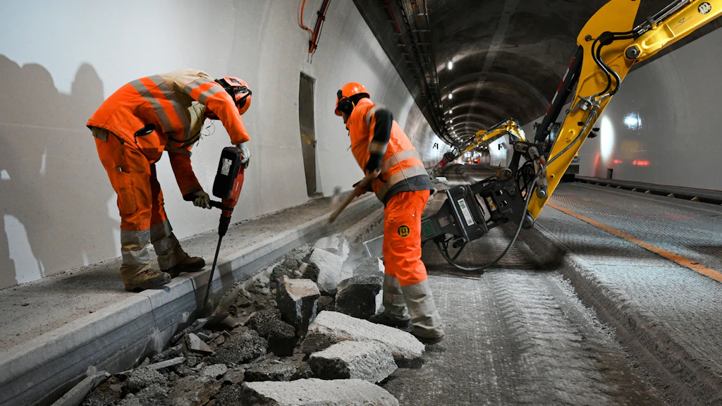 Zwei Bauarbeiter in oranger Schutzkleidung arbeiten mit Presslufthammer und Schaufel an einer aufgebrochenen Betonfahrbahn in einem Tunnel
